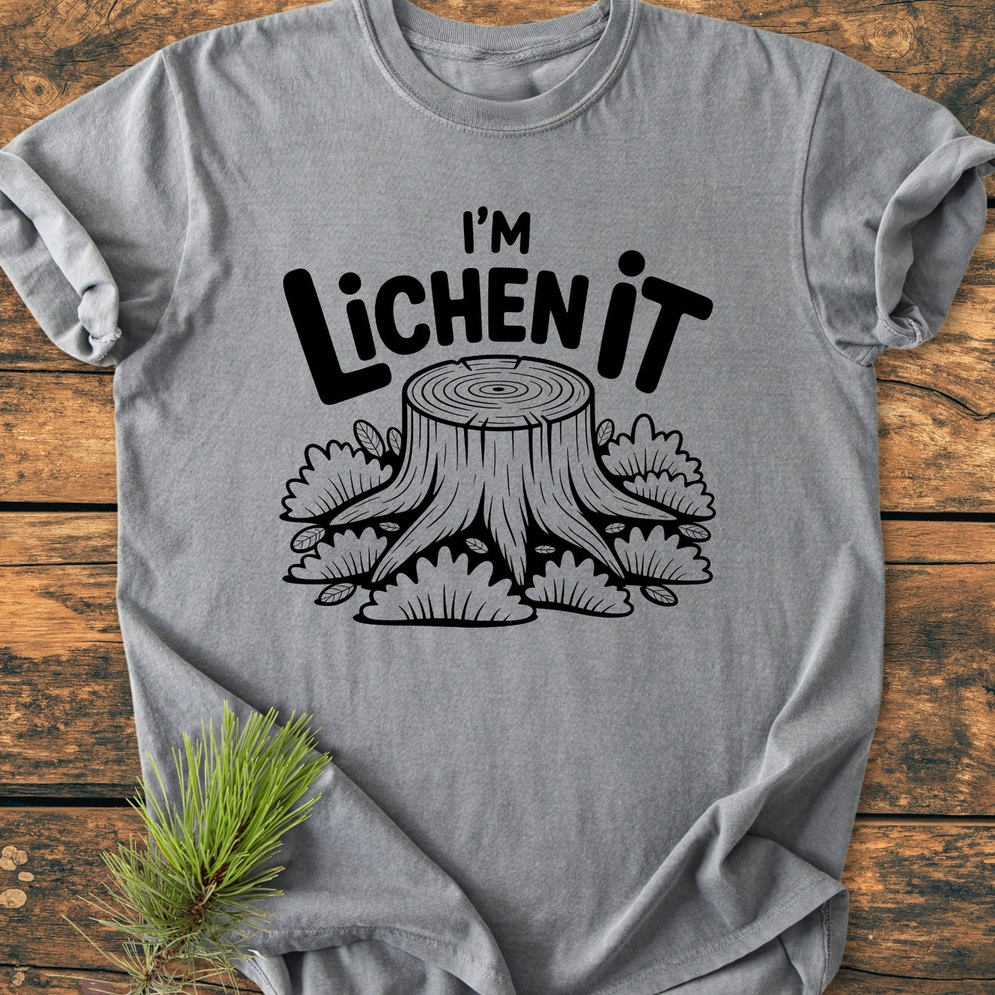 I'm Lichen It - Vintage Wash Tee on rustic wood, featuring a black tree stump illustration with moss and playful slogan. Heather-gray, relaxed fit, 100% ring-spun cotton for comfort. Pine sprig decorates scene.