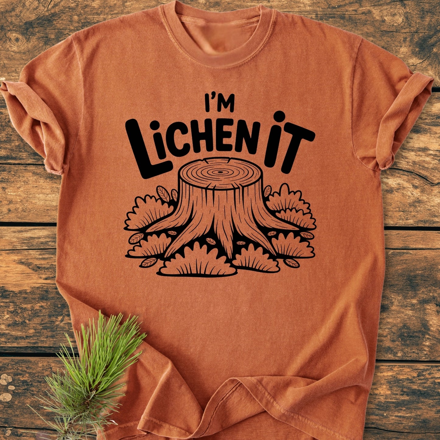 I'm Lichen It - Vintage Wash Tee features a rust-orange hue with a tree stump design and pun text, laid on wood with pine twig accenting its cozy, relaxed-fit appearance.