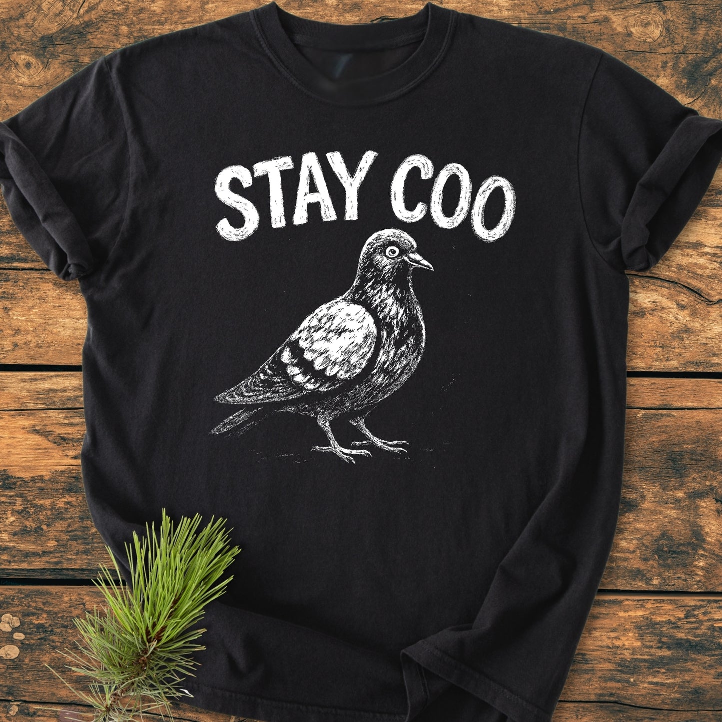 Stay Coo - Vintage Wash Tee features a white pigeon sketch with STAY COO text on a black, garment-dyed T-shirt, displayed on wood with pine needles accent, emphasizing comfort and durability.
