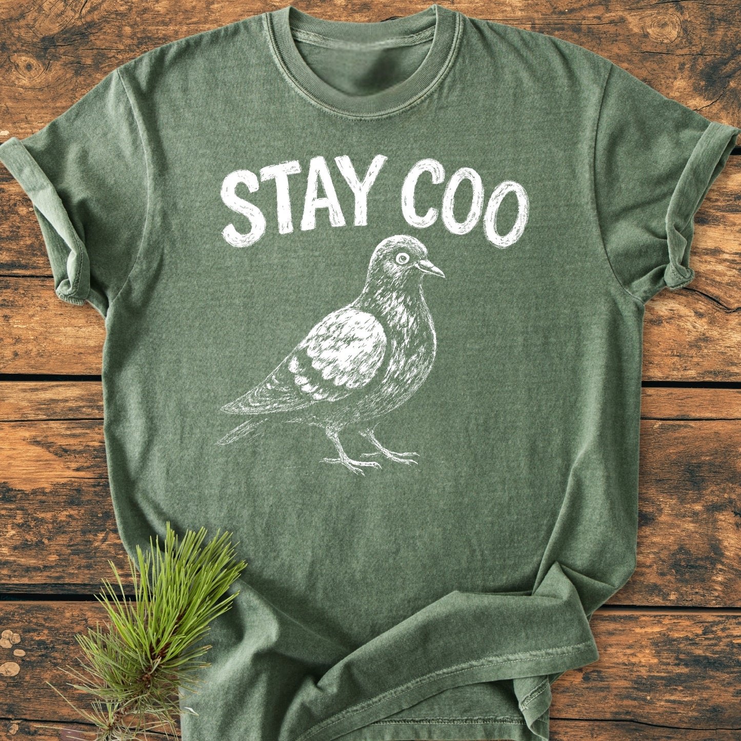 Stay Coo - Vintage Wash Tee featuring a sketch-style pigeon and STAY COO slogan, laid on a wooden surface with pine sprig. Made from soft-washed, garment-dyed 100% ring-spun cotton.