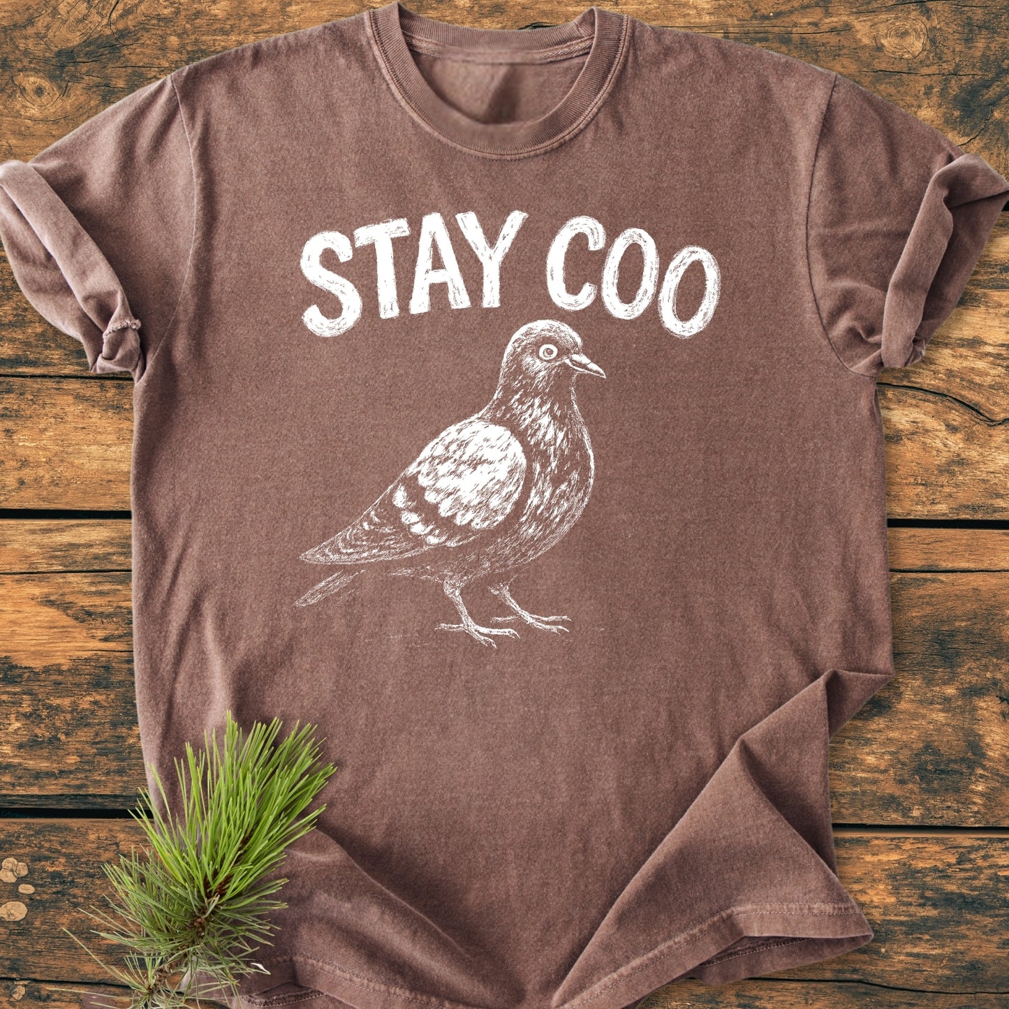 Stay Coo - Vintage Wash Tee: Heather-brown T-shirt featuring a white hand-sketch pigeon and STAY COO text, styled on rustic wood with a pine sprig, highlighting its natural, relaxed aesthetic.