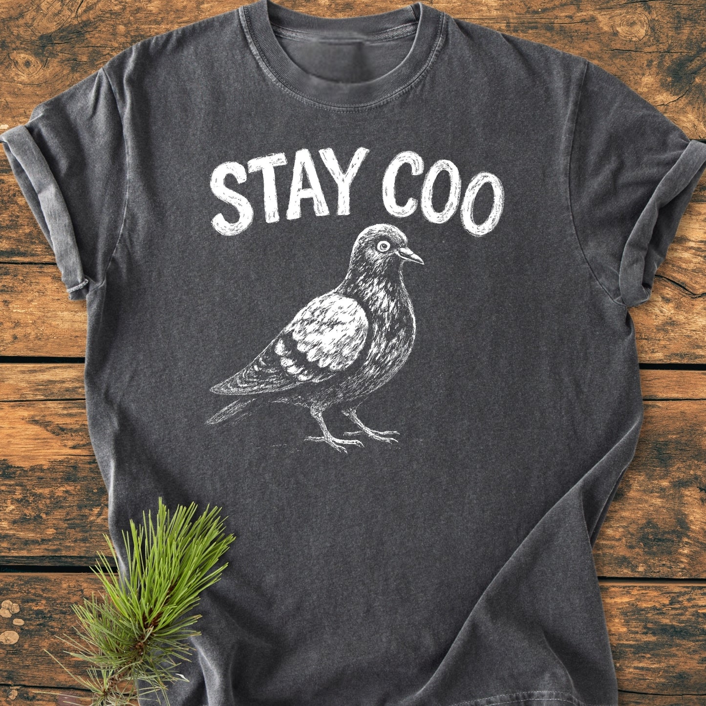 Stay Coo - Vintage Wash Tee featuring a white chalk-style pigeon illustration and STAY COO slogan on a charcoal-gray background, laid flat on wood with pine needles nearby.