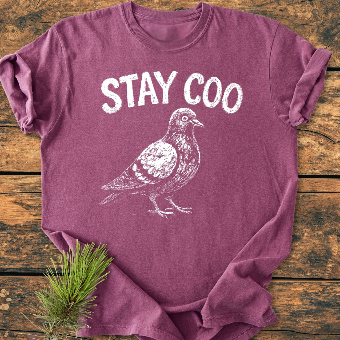 Stay Coo - Vintage Wash Tee features a chalk-style “STAY COO” print with a pigeon drawing on a mauve-purple T-shirt, laid on wood, with an evergreen sprig accent.