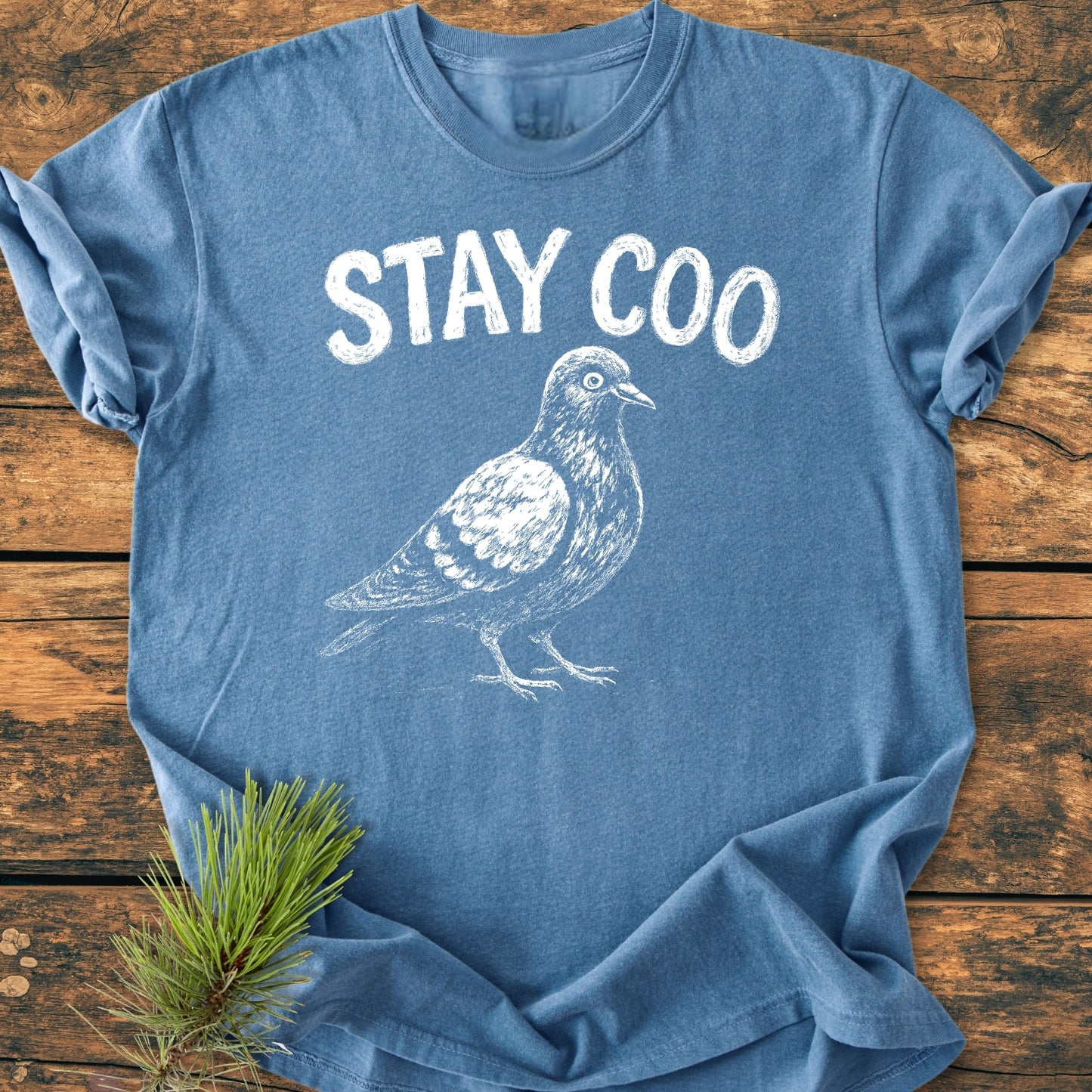 Stay Coo - Vintage Wash Tee featuring a white pigeon outline and STAY COO text on a blue, short-sleeve T-shirt, styled with pine needles on a wooden surface.