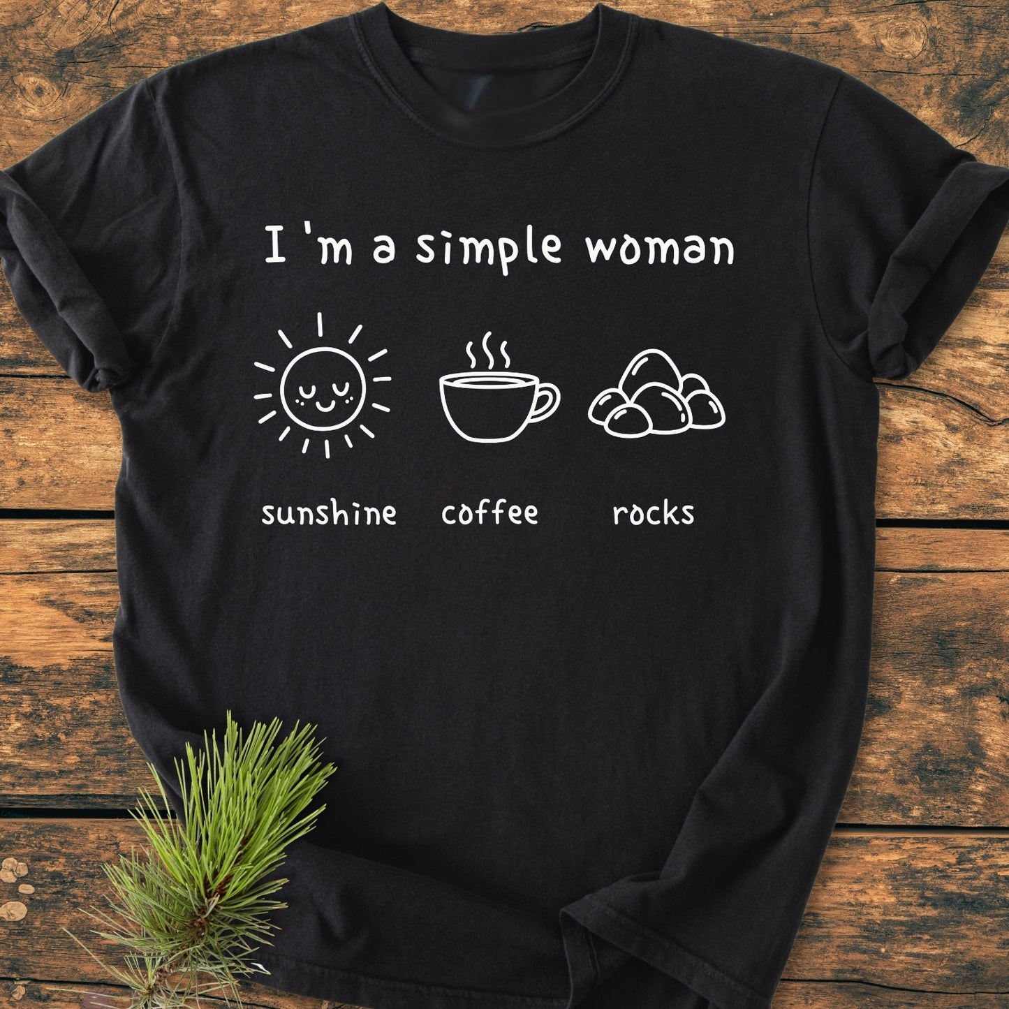 Sunshine, Coffee, Rocks - Vintage Wash Tee features a black T-shirt with “I’m a simple woman” and icons of sunshine, coffee, and rocks, laid on a wooden surface.