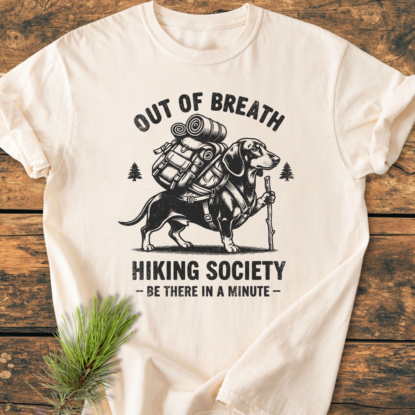 Out Of Breath Dachshund - Vintage Wash Tee: Cream T-shirt featuring a dachshund with hiking gear and humorous text. Laid on wood with a pine sprig, showcasing soft, durable Comfort Colors fabric.