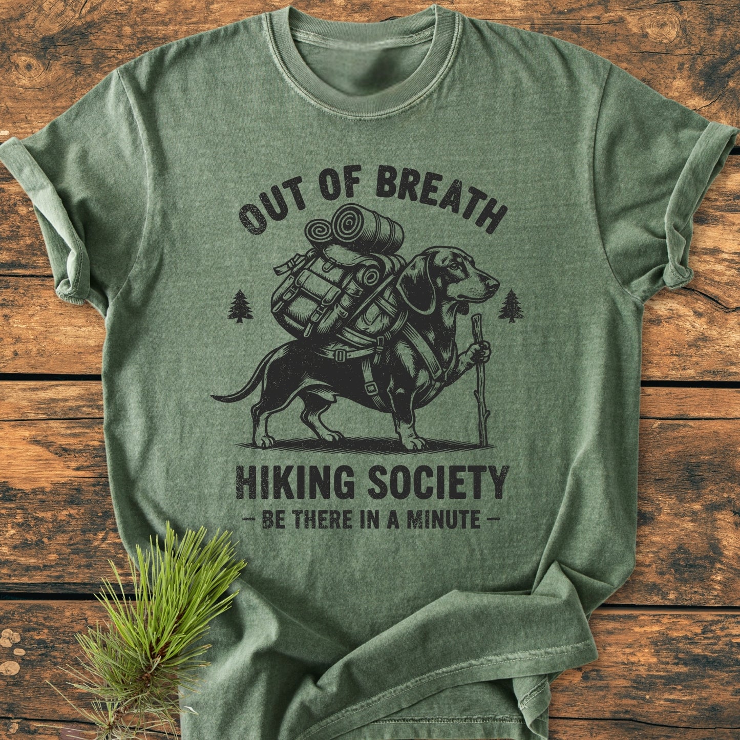 Out Of Breath Dachshund - Vintage Wash Tee features a cartoon dachshund with hiking gear, Hiking Society text, and pine-tree icons, screen-printed on a heather-green garment-dyed T-shirt.