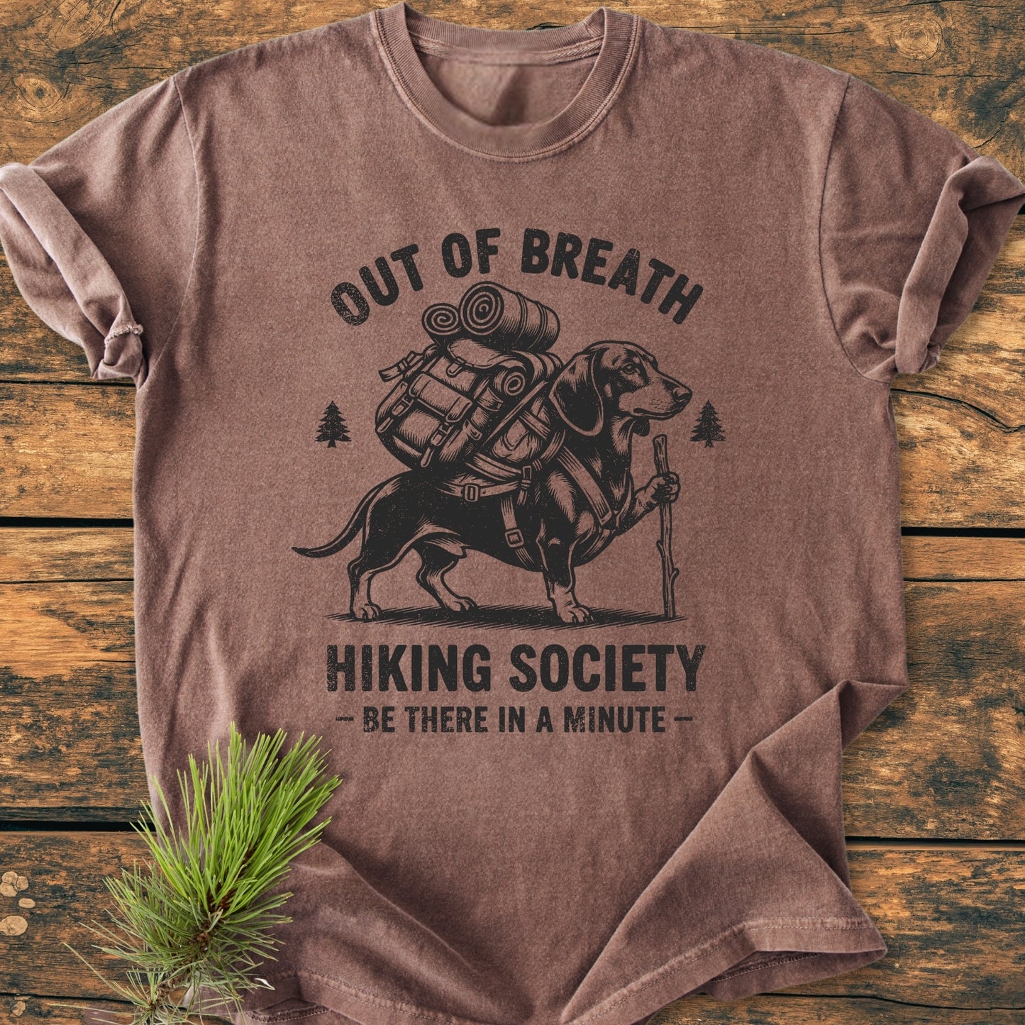 Out Of Breath Dachshund - Vintage Wash Tee featuring a dachshund with hiking gear, printed on a brown, short-sleeved T-shirt, laid on a wooden surface with a pine branch nearby.