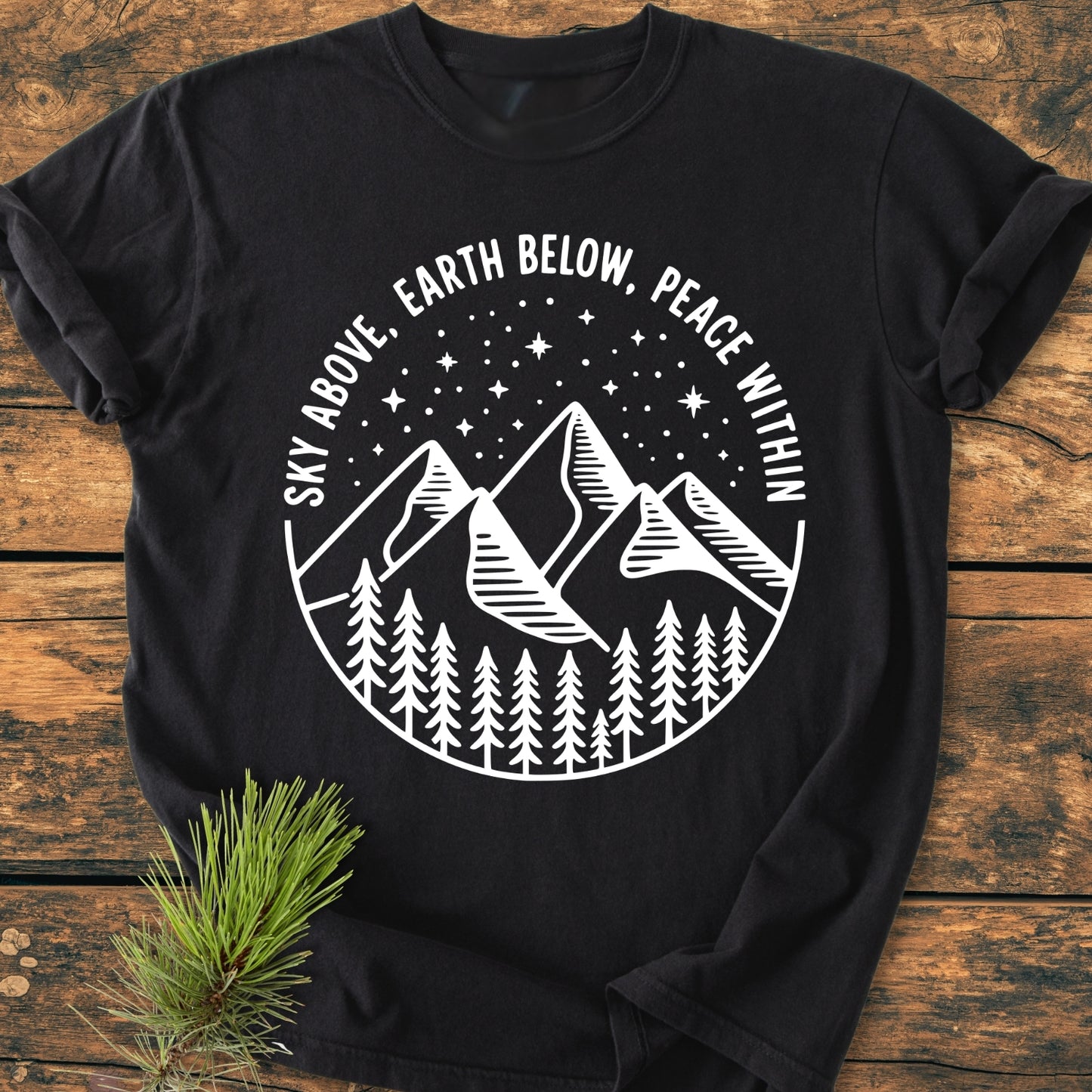 Sky Above - Vintage Wash Tee featuring a circular design with pine trees, mountains, and a starry sky. Motto reads: Sky Above, Earth Below, Peace Within. Laid on wooden surface with evergreen sprig.