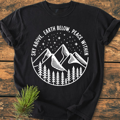 Sky Above - Vintage Wash Tee featuring a circular design with pine trees, mountains, and a starry sky. Motto reads: Sky Above, Earth Below, Peace Within. Laid on wooden surface with evergreen sprig.