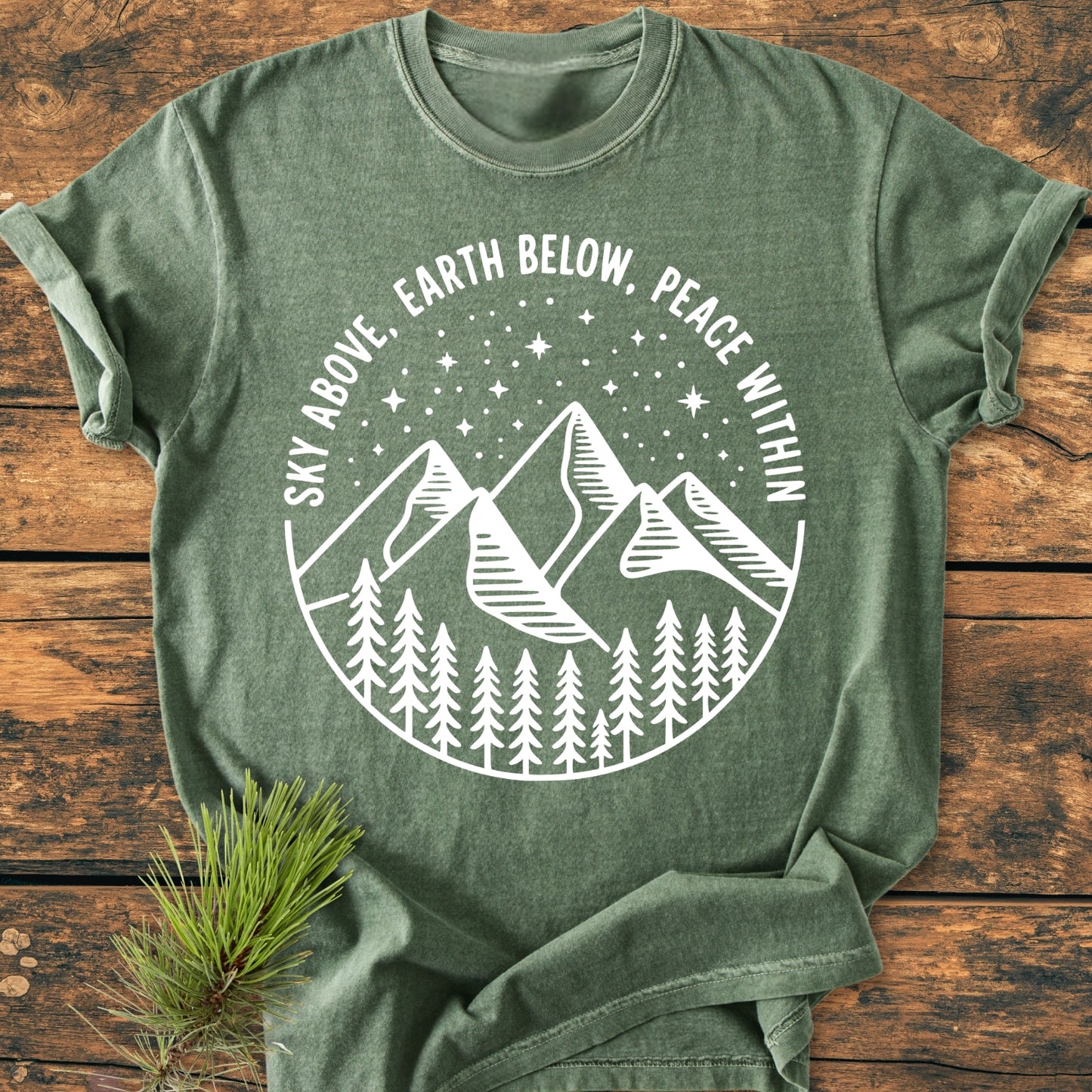 Sky Above - Vintage Wash Tee on wooden surface, features a circular graphic with pine trees, mountains, and a starry sky, accented by the motto “SKY ABOVE, EARTH BELOW, PEACE WITHIN.” Pine sprig beside.