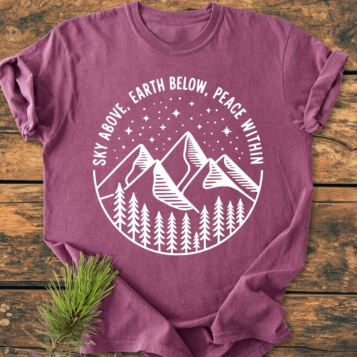 Sky Above - Vintage Wash Tee, featuring a white circular graphic with mountains, pine trees, and stars, laid on wood. The heather-purple tee offers a relaxed fit with durable double-needle stitching.
