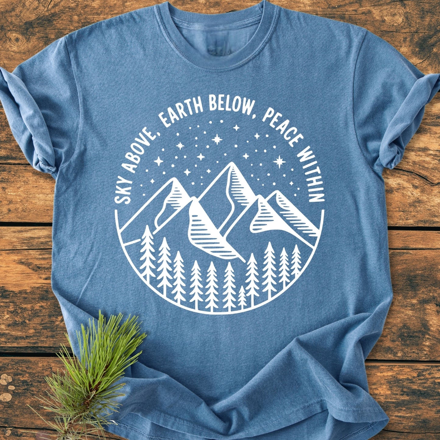 Sky Above - Vintage Wash Tee featuring a white circular design of pine trees, mountains, and stars on heather-blue fabric, highlighting the phrase “SKY ABOVE, EARTH BELOW, PEACE WITHIN.”