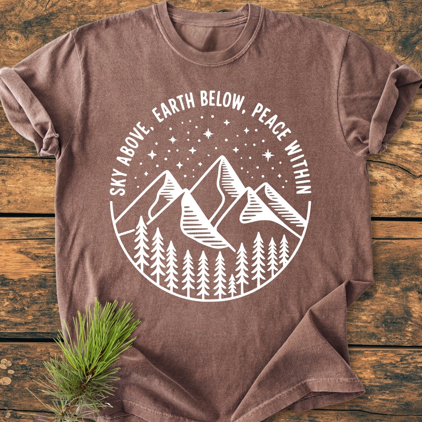 Sky Above - Vintage Wash Tee featuring a white line-art design of mountains and pine trees under a starry sky on a brown T-shirt, laid on a wooden surface with a pine sprig beside it.
