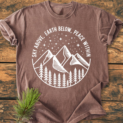 Sky Above - Vintage Wash Tee featuring a white line-art design of mountains and pine trees under a starry sky on a brown T-shirt, laid on a wooden surface with a pine sprig beside it.
