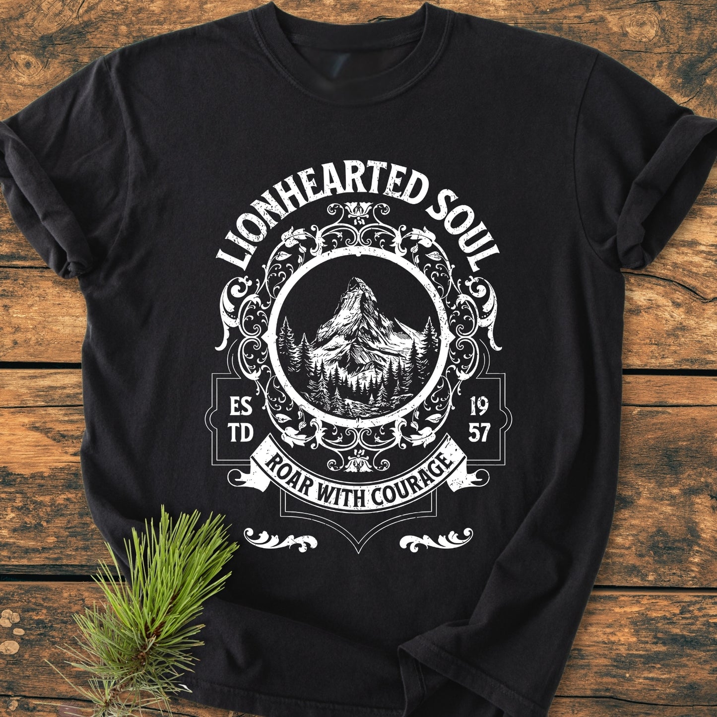 Lionhearted Soul - Vintage Wash Tee features a white, vintage-style emblem with mountains and pine trees on a black T-shirt, laid flat on a rustic wood surface with a nearby pine sprig.