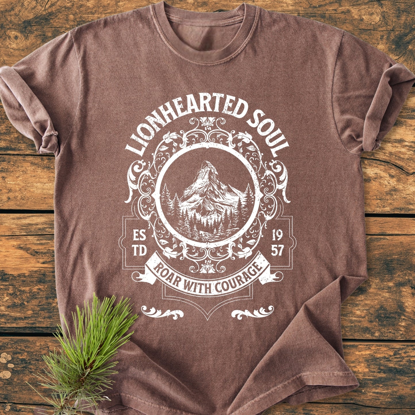 Lionhearted Soul - Vintage Wash Tee features a brown, vintage-washed look with a white ornate crest of a mountain and forest scene, emphasizing ROAR WITH COURAGE, laid flat on a wooden surface.