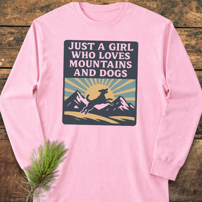 Just A Girl Who Loves Dogs Comfort Colors Long-Sleeve Tee featuring a rising sun, mountains, and dog silhouette graphic on soft fabric, laid on a wooden surface with a pine sprig beside.