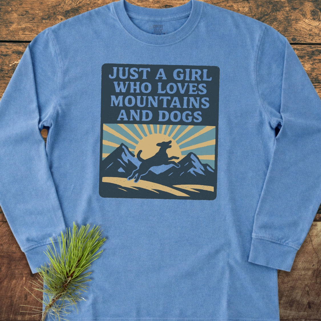 Just A Girl Who Loves Dogs Comfort Colors Long-Sleeve Tee featuring a mountain and dog graphic with text, laid out on a wooden surface beside a pine sprig, emphasizing its casual, relaxed style.