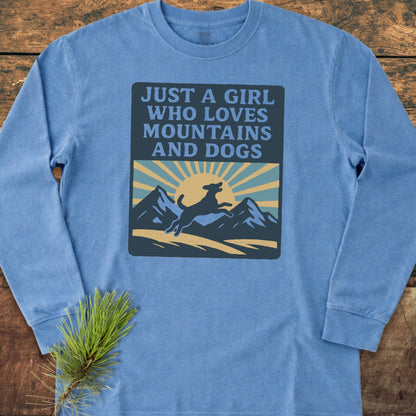Just A Girl Who Loves Dogs Comfort Colors Long-Sleeve Tee featuring a mountain and dog graphic with text, laid out on a wooden surface beside a pine sprig, emphasizing its casual, relaxed style.