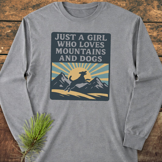 Just A Girl Who Loves Dogs Comfort Colors Long-Sleeve Tee featuring a mountain and dog graphic with a rising sun, laid flat on wood, highlighting its 100% ring-spun cotton softness and relaxed fit.
