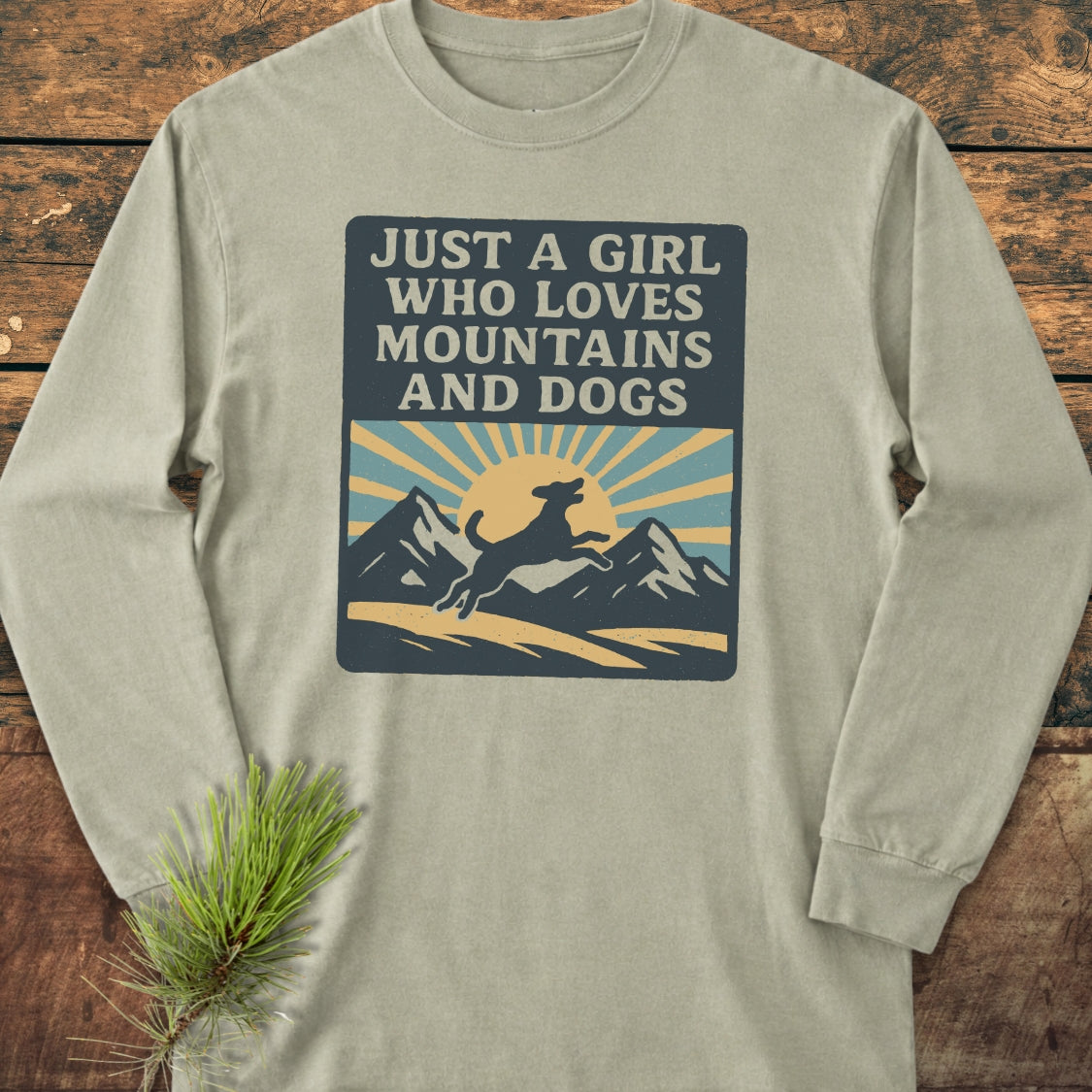 Just A Girl Who Loves Dogs Comfort Colors Long-Sleeve Tee features a bold graphic with mountains, sun, and a leaping dog, laid flat on wood with a pine sprig accent.