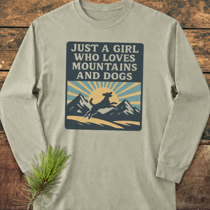 Just A Girl Who Loves Dogs Comfort Colors Long-Sleeve Tee features a bold graphic with mountains, sun, and a leaping dog, laid flat on wood with a pine sprig accent.