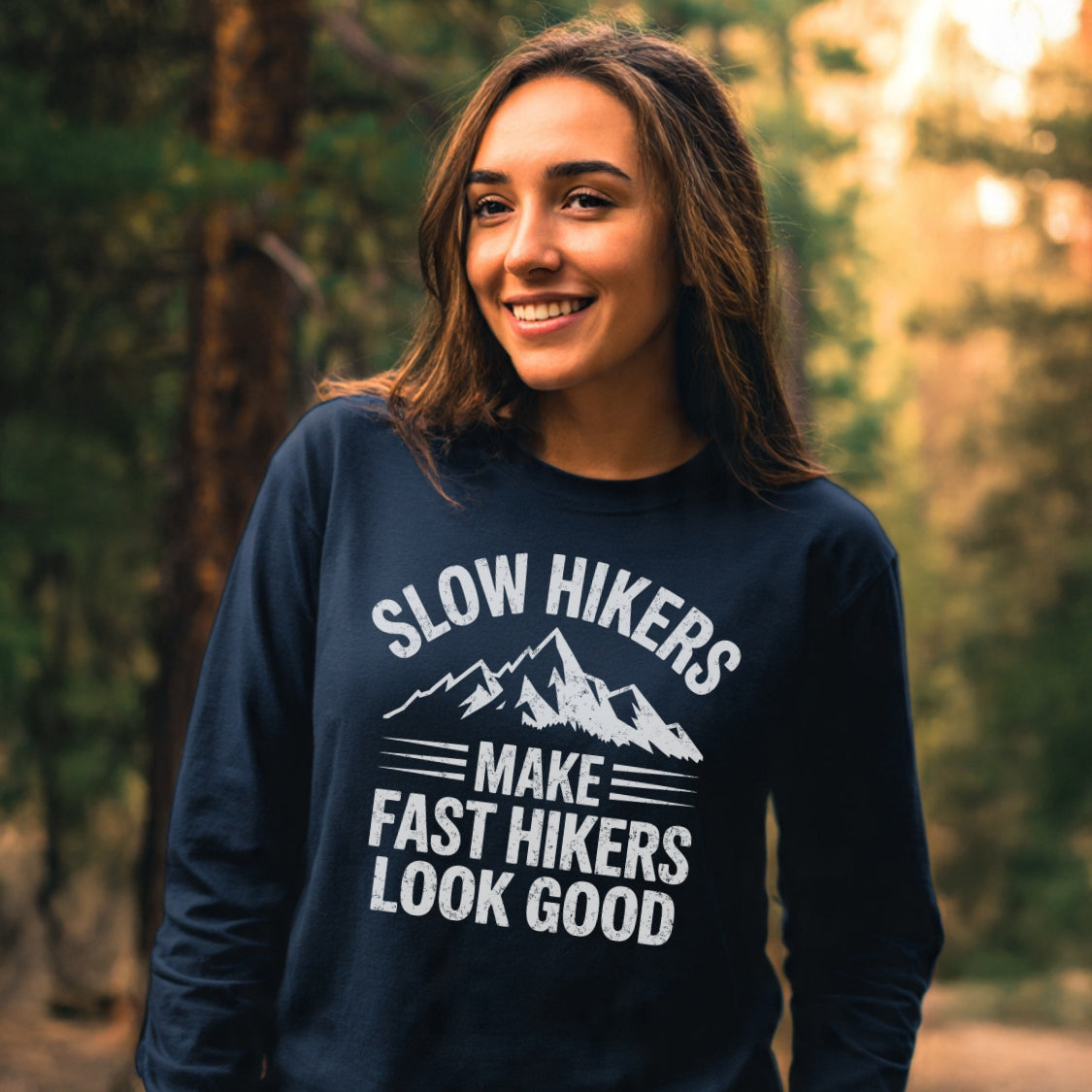 Young woman in a forest wearing the Slow Hikers Make Fast Hikers Look Good Long-Sleeve Tee, showcasing a relaxed fit and mountain graphic, perfect for casual, comfortable outings.