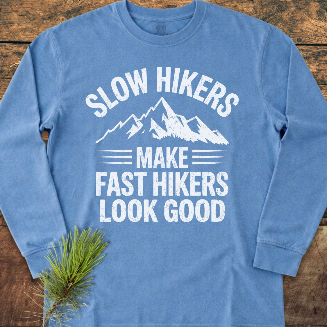 Slow Hikers Make Fast Hikers Look Good Long-Sleeve Tee displayed on a wooden surface, featuring a white mountain graphic and slogan, with a pine sprig accentuating its outdoorsy appeal.