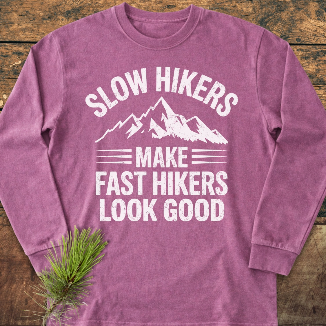Slow Hikers Make Fast Hikers Look Good Long-Sleeve Tee displayed on wooden surface, featuring white mountain graphic and slogan, crafted from 100% ring-spun cotton for softness and a relaxed fit.