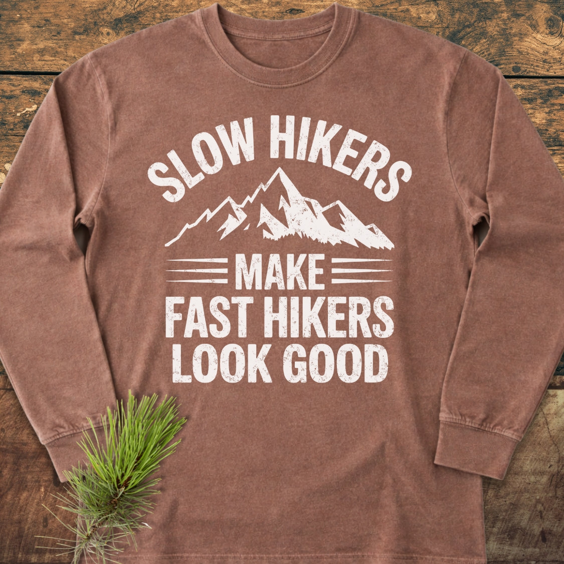 Slow Hikers Make Fast Hikers Look Good Long-Sleeve Tee, featuring a white mountain graphic and slogan on a brown shirt, laid on wood with a pine sprig, emphasizing comfort and style.
