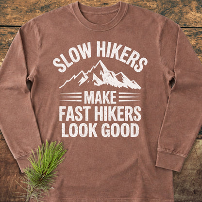 Slow Hikers Make Fast Hikers Look Good Long-Sleeve Tee, featuring a white mountain graphic and slogan on a brown shirt, laid on wood with a pine sprig, emphasizing comfort and style.