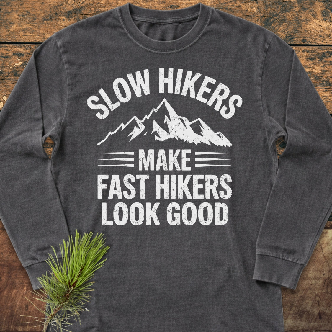 Slow Hikers Make Fast Hikers Look Good Long-Sleeve Tee featuring a mountain graphic and slogan on a wooden surface with a pine sprig accentuating its outdoorsy appeal.