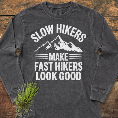Slow Hikers Make Fast Hikers Look Good Long-Sleeve Tee featuring a mountain graphic and slogan on a wooden surface with a pine sprig accentuating its outdoorsy appeal.