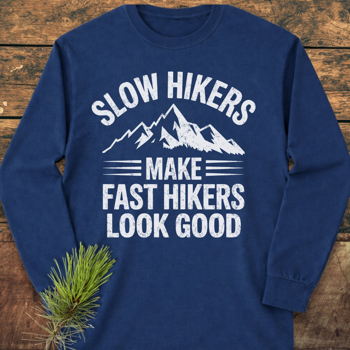 Navy-blue Slow Hikers Make Fast Hikers Look Good Long-Sleeve Tee with a distressed mountain graphic and humorous text, laid flat on wood, featuring a relaxed fit for casual comfort.