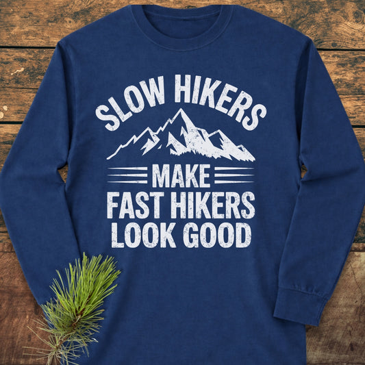 Navy-blue Slow Hikers Make Fast Hikers Look Good Long-Sleeve Tee with a distressed mountain graphic and humorous text, laid flat on wood, featuring a relaxed fit for casual comfort.