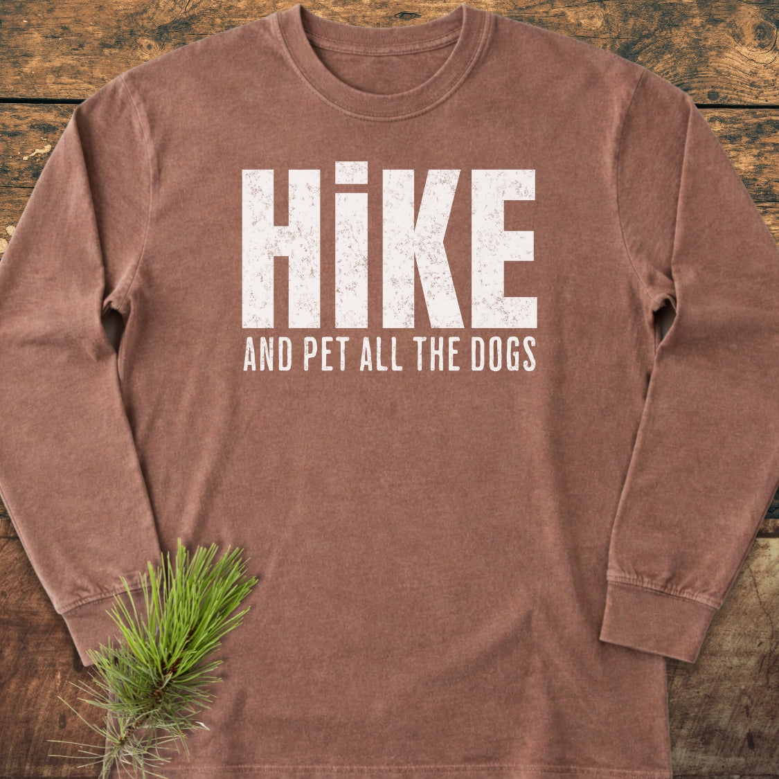 Hike And Pet Dogs Long-Sleeve Tee displayed on wood, featuring bold white distressed HIKE text with AND PET ALL THE DOGS below; includes a pine sprig accent, emphasizing its relaxed, comfortable style.