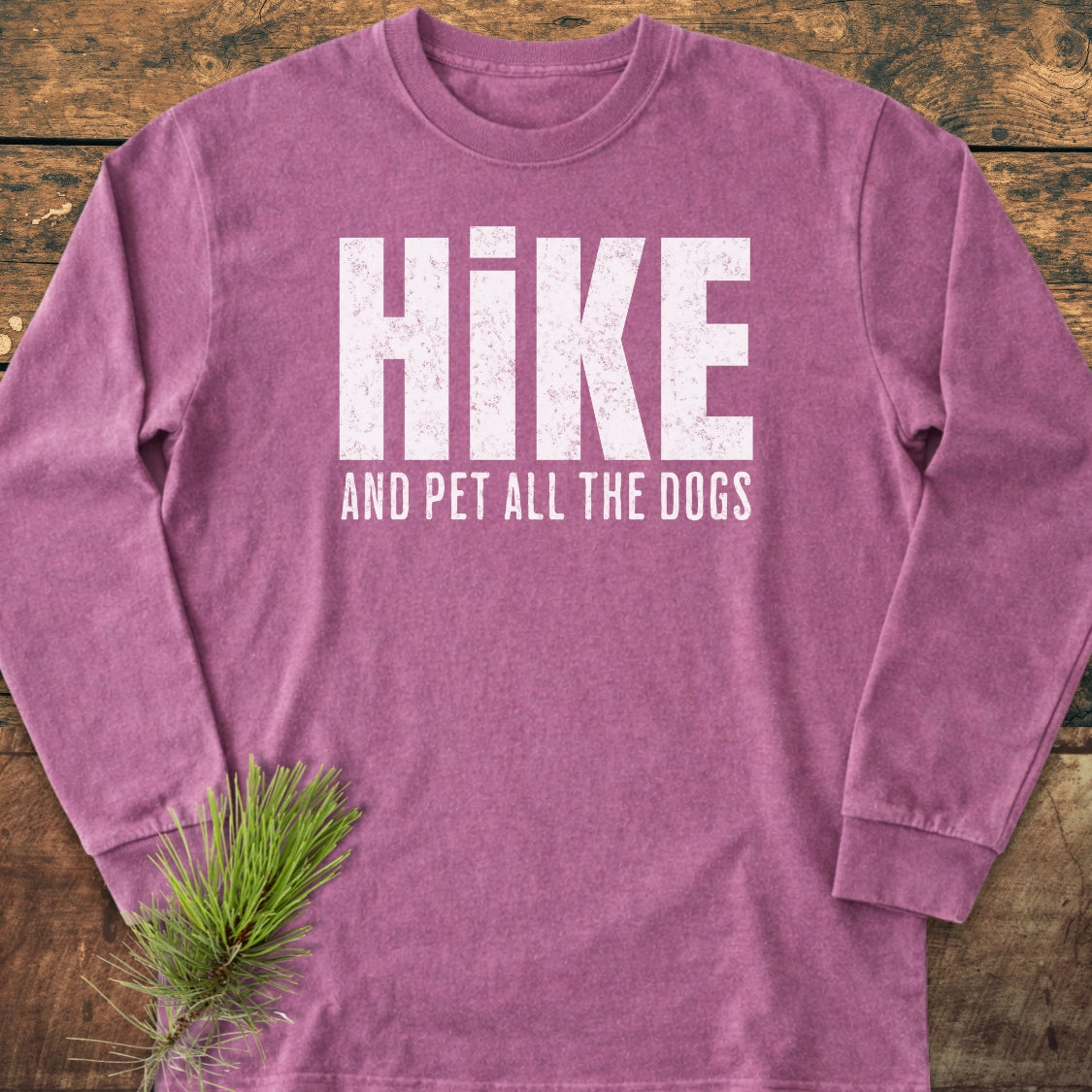 Hike And Pet Dogs Long-Sleeve Tee, heathered purple, features distressed white lettering on the chest, laid on a wooden surface with a pine sprig. Made from 100% ring-spun cotton for a relaxed fit.