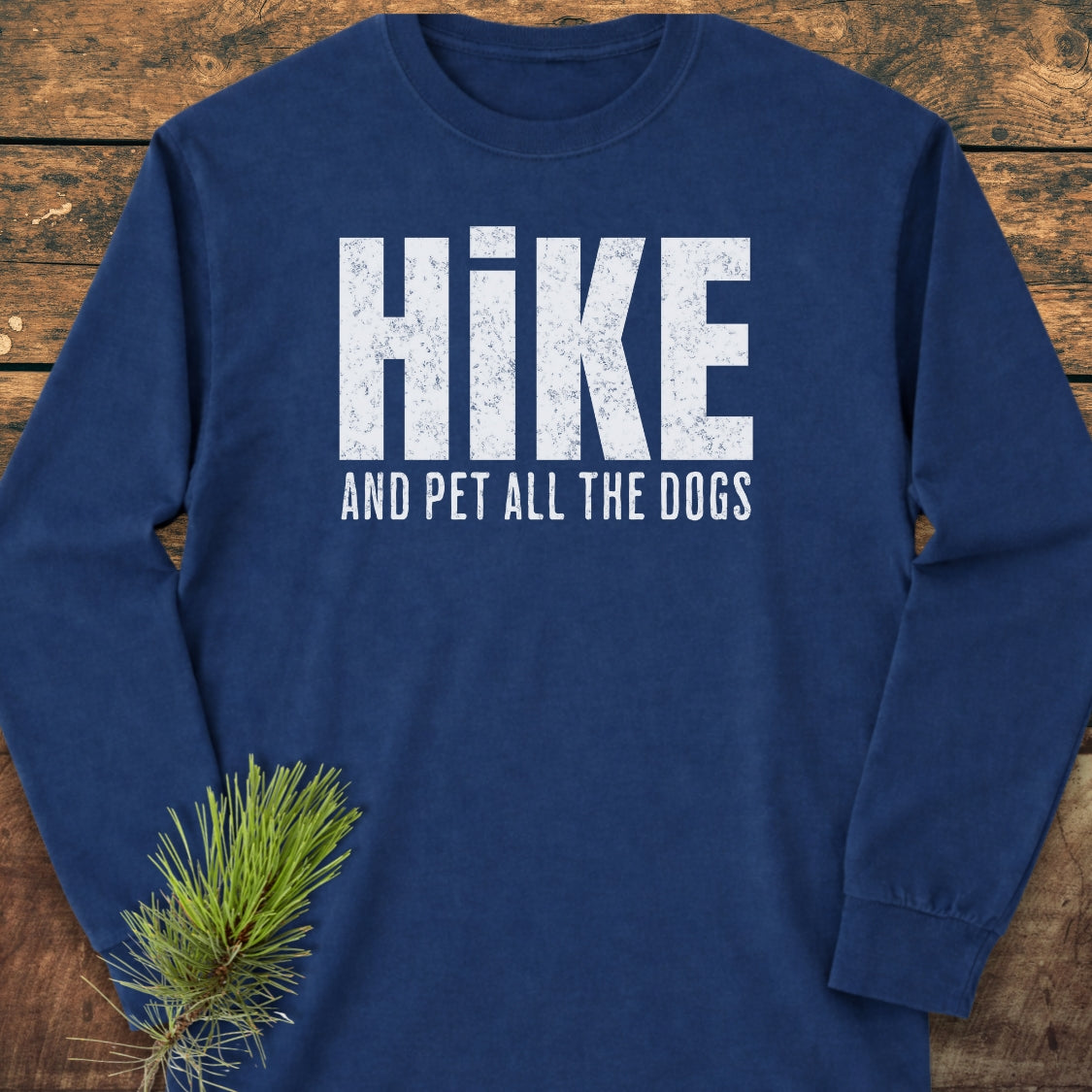 Hike And Pet Dogs Long-Sleeve Tee, featuring distressed white slogan on navy-blue, laid on wood. Includes pine sprig accent. Made from 100% ring-spun cotton with relaxed fit for comfort.