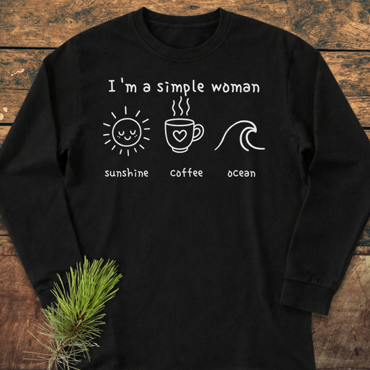 I'm A Simple Woman - Sunshine Coffee Ocean Long-Sleeve Tee features black fabric with white graphics of a sun, coffee mug, and wave. Laid on wood, accented by a pine sprig.