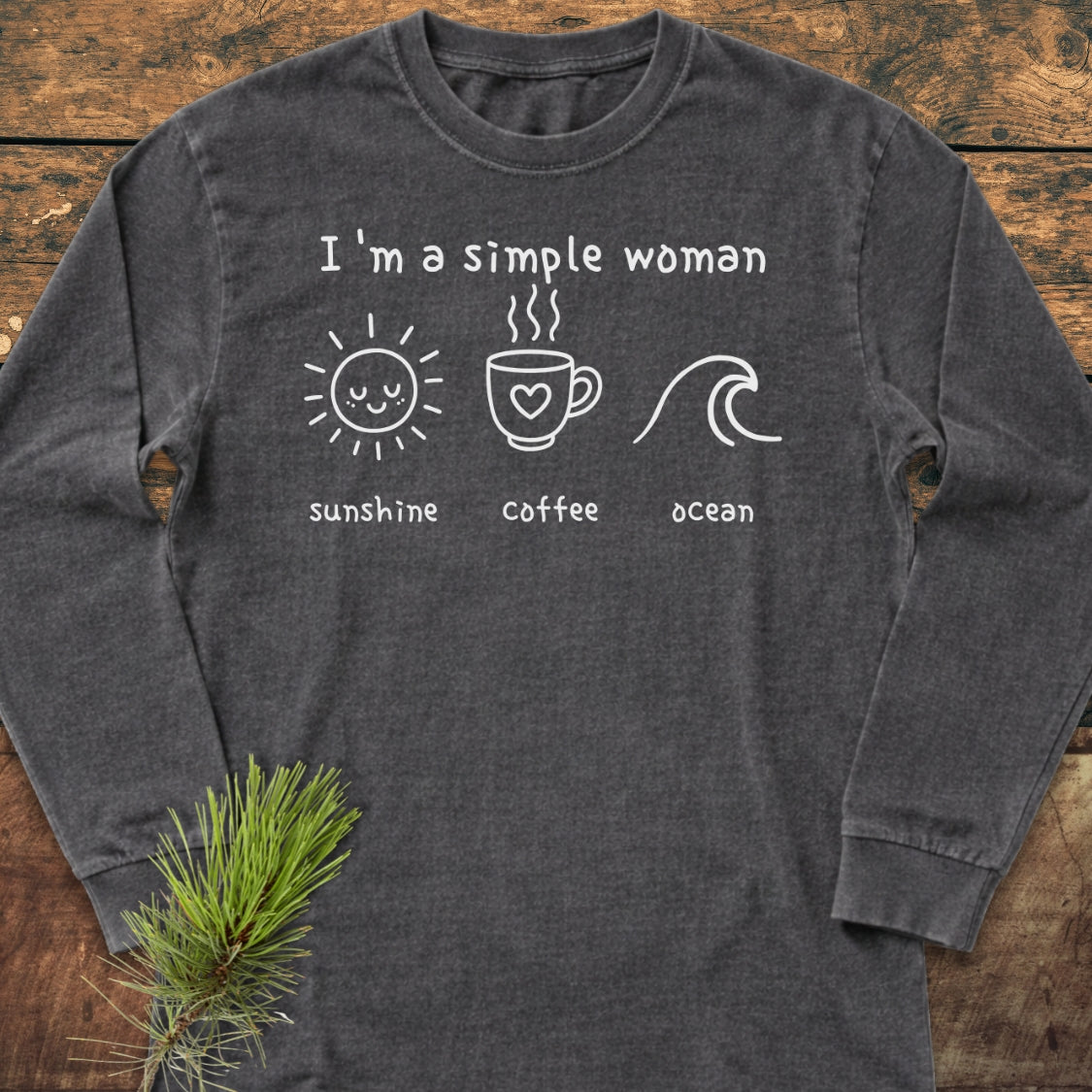 I'm A Simple Woman - Sunshine Coffee Ocean Long-Sleeve Tee featuring hand-written text and line drawings of a sun, coffee cup, and wave on charcoal-gray fabric, displayed on a wooden surface.