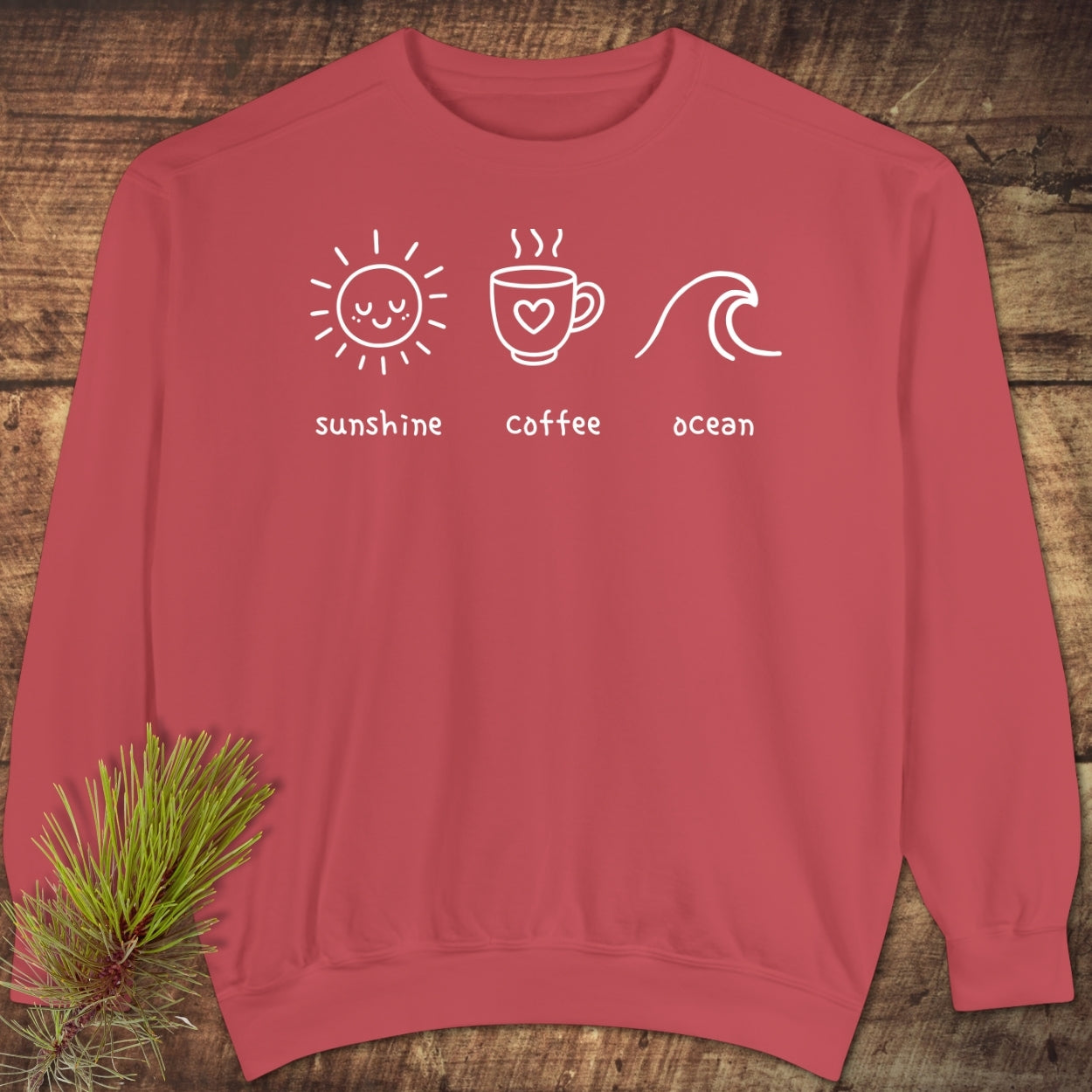 I'm A Simple Woman - Sunshine Coffee Ocean Sweatshirt featuring a coral crewneck design with white-line illustrations of a sun, coffee mug, and ocean wave, laid flat on a wooden surface.