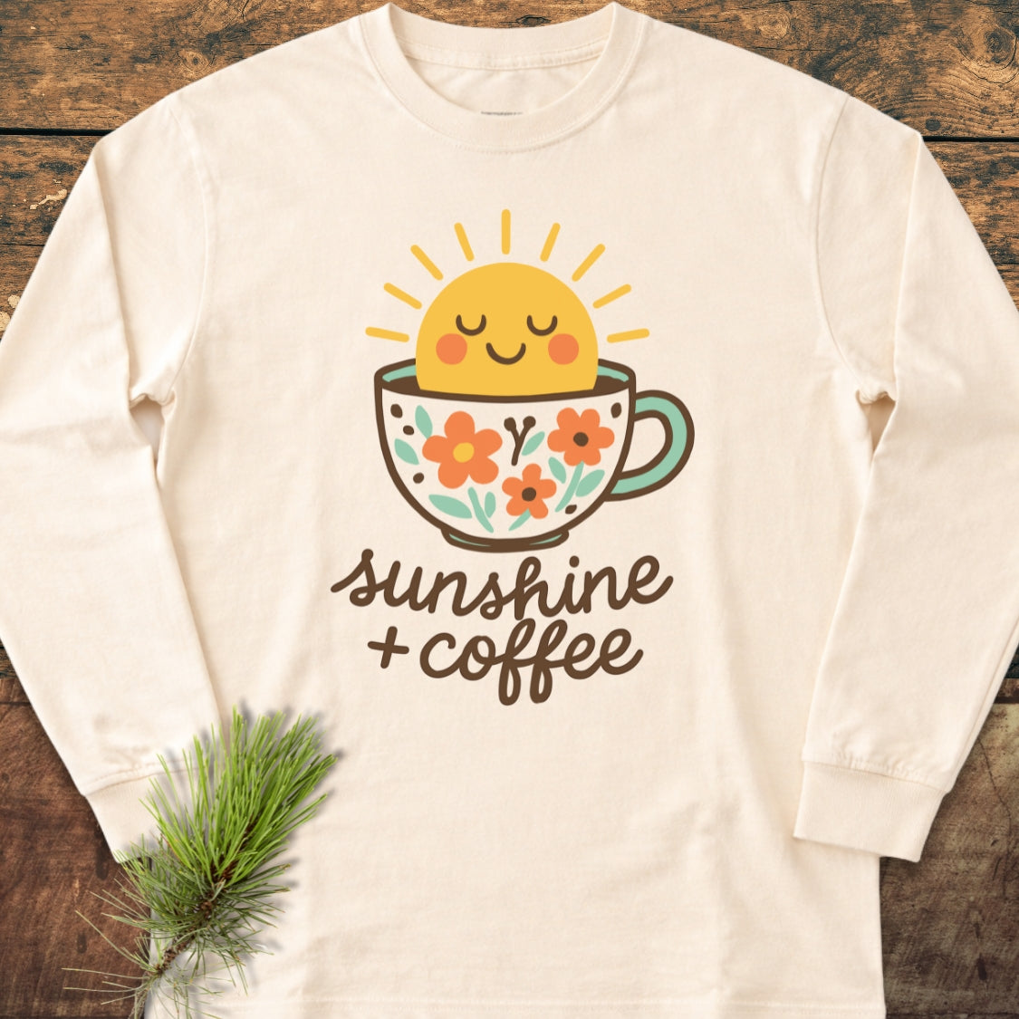 Sunshine And Coffee Long-Sleeve Tee featuring a smiling sun in a floral coffee cup with sunshine + coffee text, laid on wood with a pine sprig, showcasing its soft, relaxed-fit cotton design.