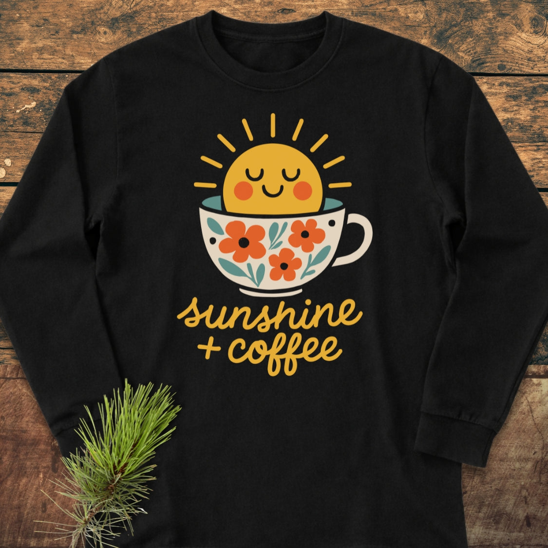 Sunshine And Coffee Long-Sleeve Tee featuring a stylized coffee cup with orange flowers, green leaves, and a smiling sun. Laid flat on wood, includes sunshine + coffee text. Made from 100% ring-spun cotton.