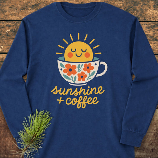 Sunshine And Coffee Long-Sleeve Tee featuring a smiling sun emerging from a floral coffee cup graphic, laid on wood with a pine sprig, offers a relaxed fit and soft, 100% cotton fabric.