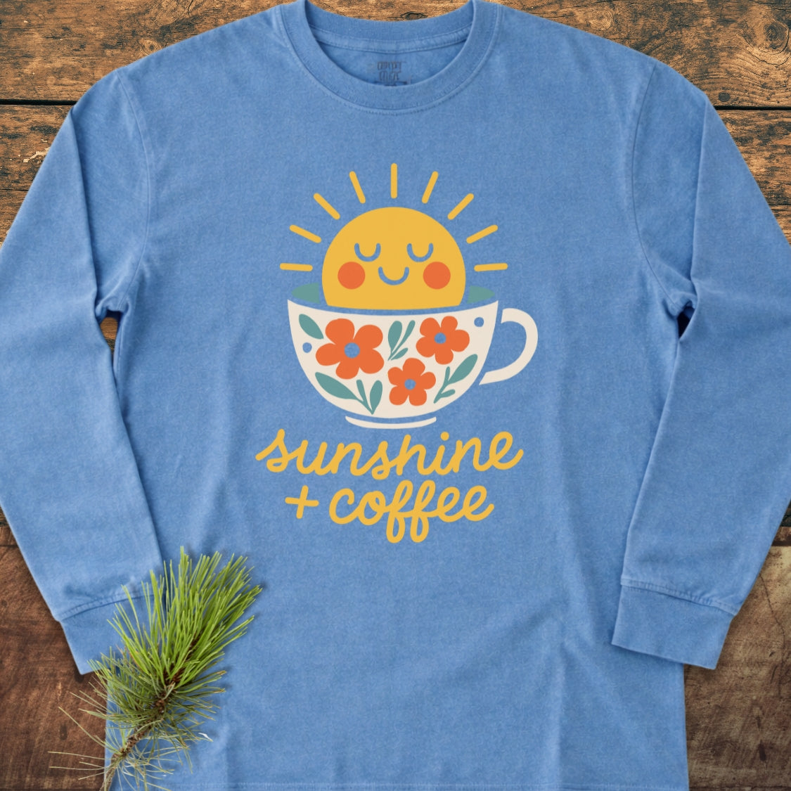 Sunshine And Coffee Long-Sleeve Tee featuring a smiling sun in a coffee cup with floral accents, laid flat with a pine sprig. Made of soft, 100% ring-spun cotton for a relaxed fit.