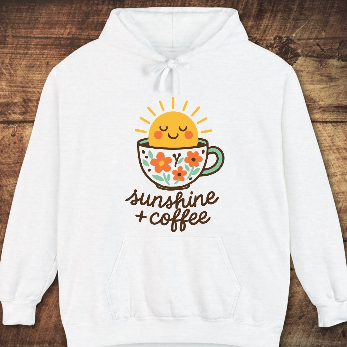 Sunshine And Coffee Hoodie with a colorful sun and coffee cup graphic, featuring sunshine + coffee text. Laid on wood surface, showcasing its cozy, garment-dyed fabric and relaxed unisex fit.