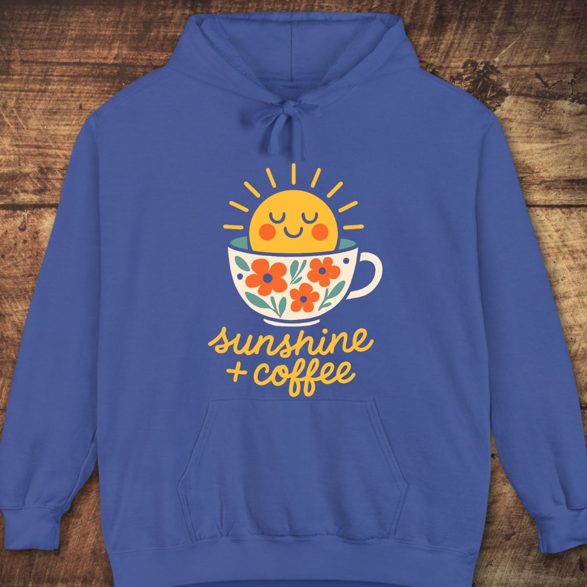 Sunshine And Coffee Hoodie featuring a smiling sun in a coffee cup with floral accents, hand-lettered script, laid flat on a wooden backdrop, showcasing its cozy, relaxed unisex fit.