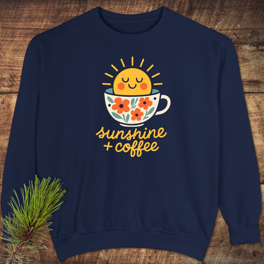 Sunshine And Coffee Sweatshirt displayed on wood, featuring a sun rising from a floral coffee cup with sunshine + coffee text. Styled with a pine sprig, showcasing its cozy, relaxed fit.