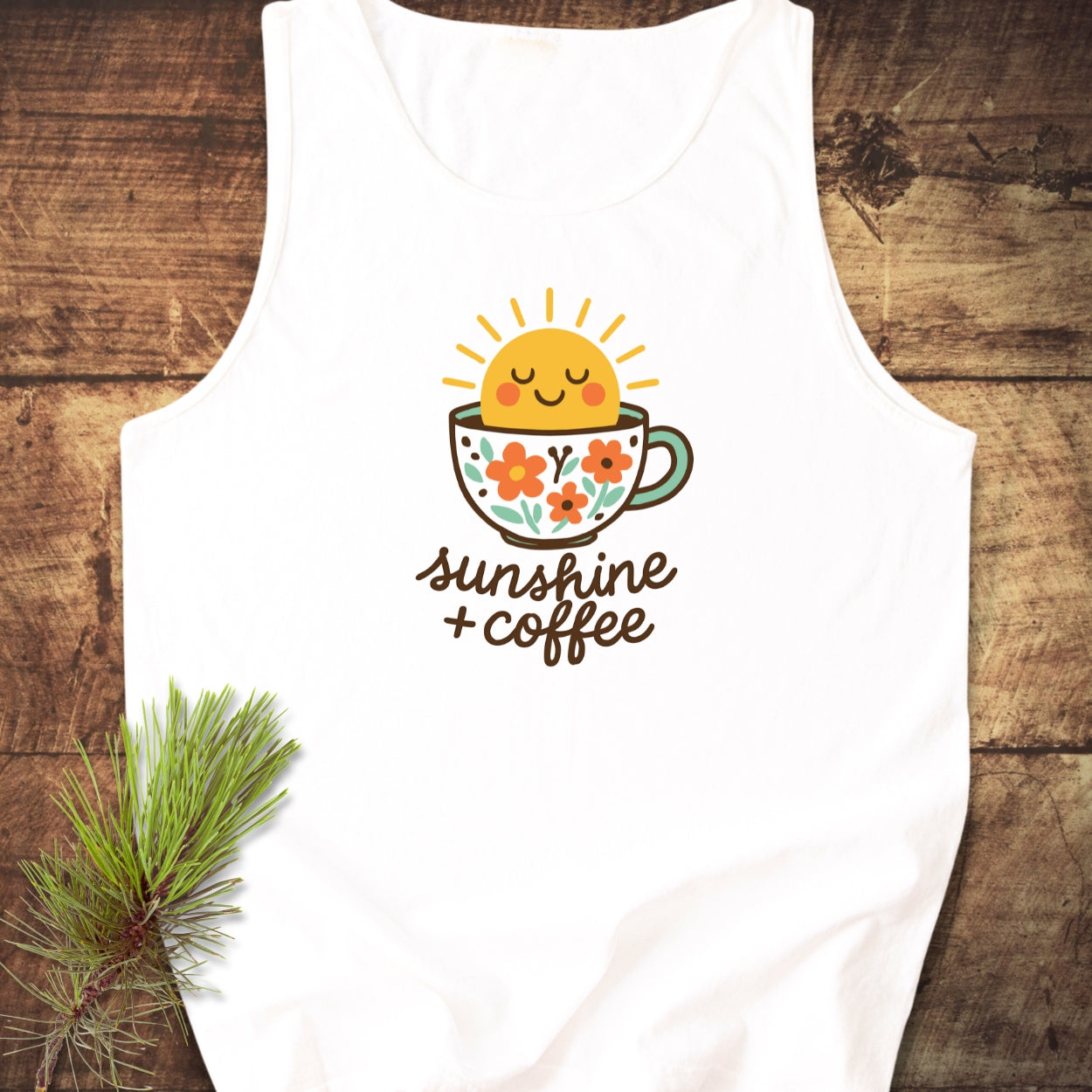 Sunshine And Coffee Tank Top featuring a smiling sun in a floral coffee cup graphic with sunshine + coffee text, displayed flat on wood with a pine branch accent.